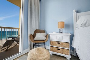 Ein Schlafzimmer mit Blick auf den Strand, ein Korbstuhl, ein Nachttisch mit Lampe und ein Bett mit weißer Bettwäsche.