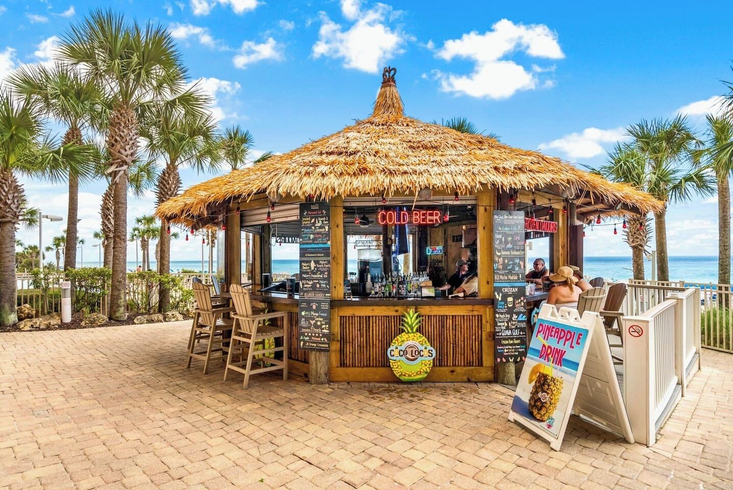 A beachside kiosk with a thatched roof, offering cold beer and pineapple drinks, with a view of the ocean.