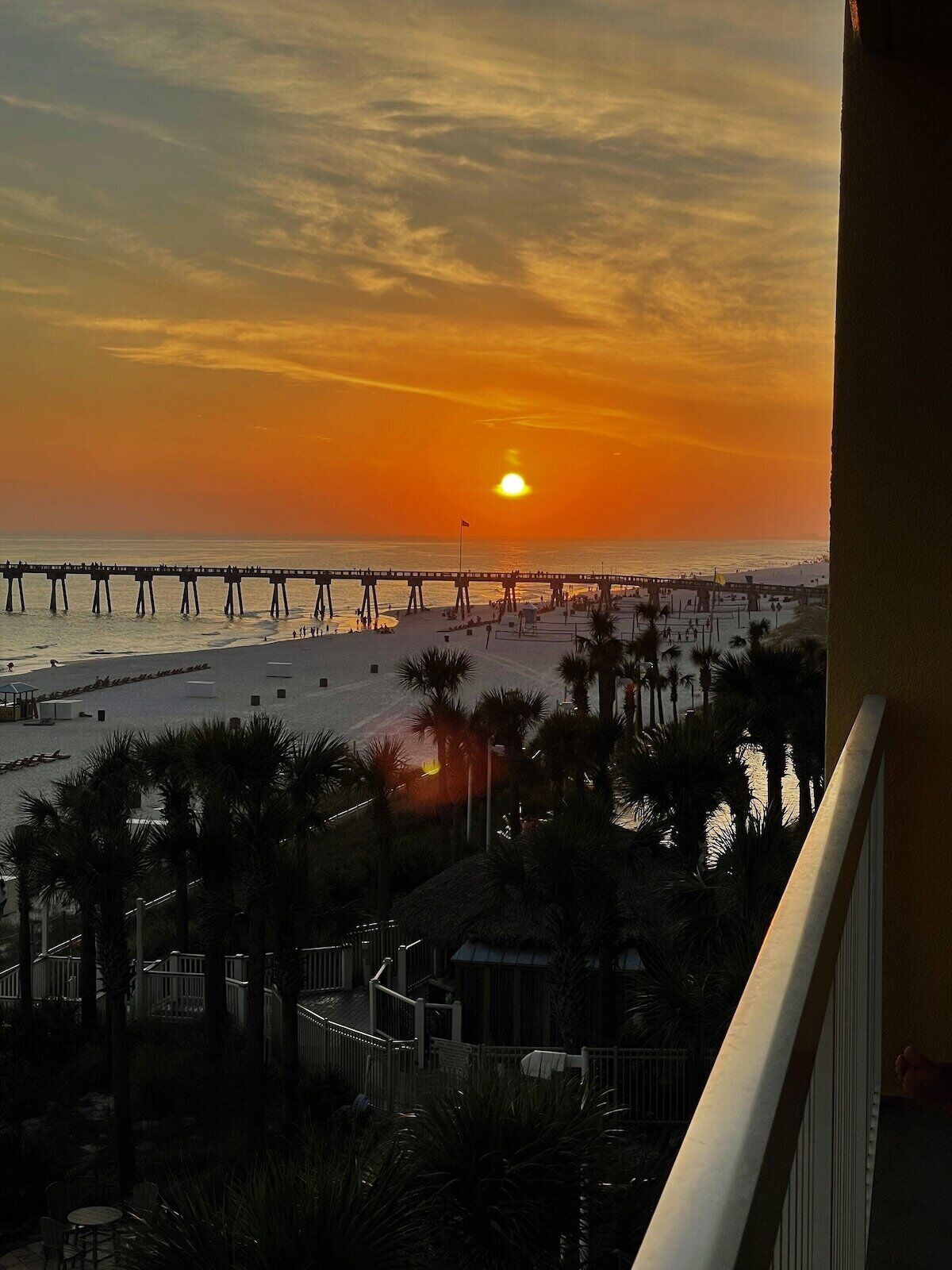Une vue sur le coucher de soleil depuis un balcon donnant sur une plage, avec une jetée et des palmiers.