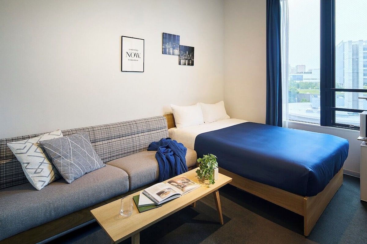 A hotel room with a bed, a sofa, a small table with a book and a glass, a microwave, and a window with curtains.