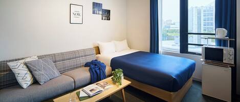 A hotel room with a bed, a sofa, a small table with a book and a glass, a microwave, and a window with curtains.
