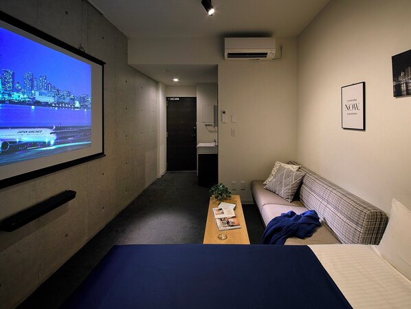 Habitación de hotel con cama, sofá, mesa de centro con una planta y un proyector montado en la pared mostrando un paisaje urbano.