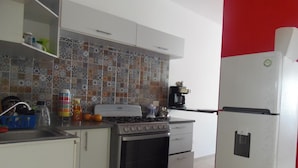 A kitchen with a stove, oven, and refrigerator. There is a tiled backsplash and a shelf with various items.
