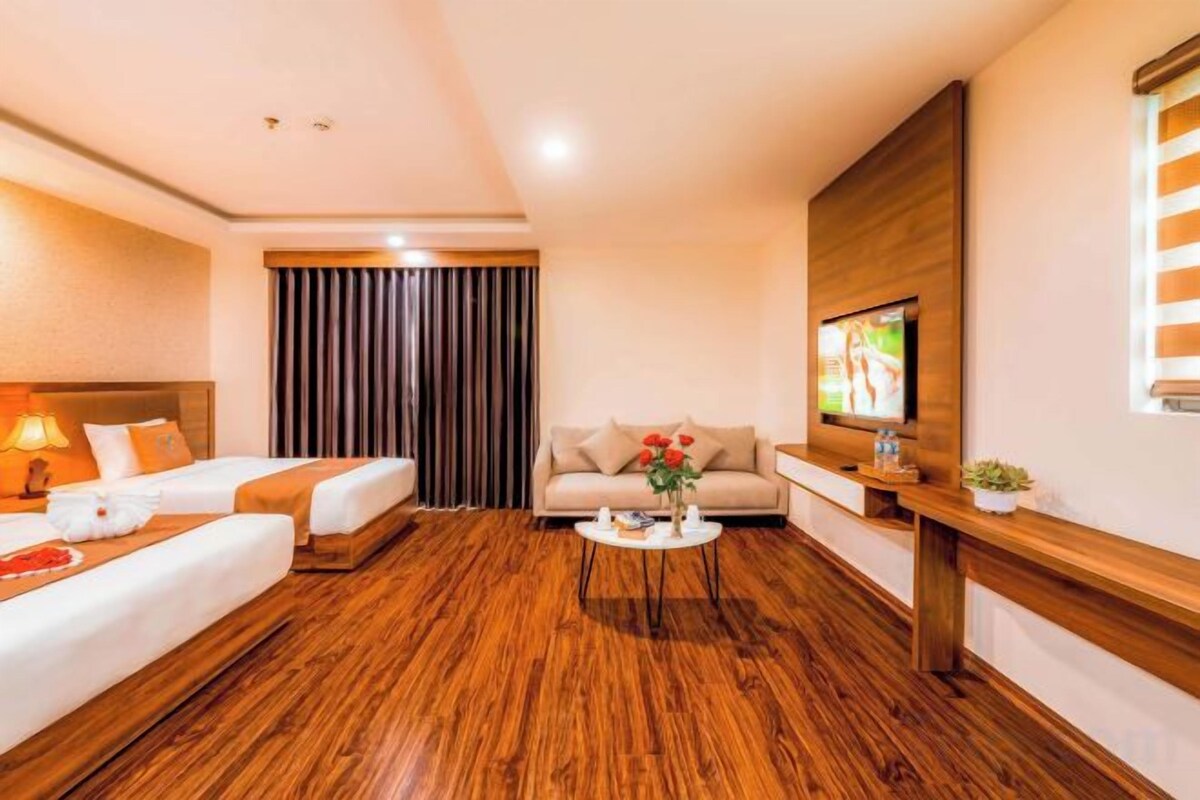 A hotel room with a wooden floor, a bed, a sofa, a coffee table, a TV, and a window with curtains.
