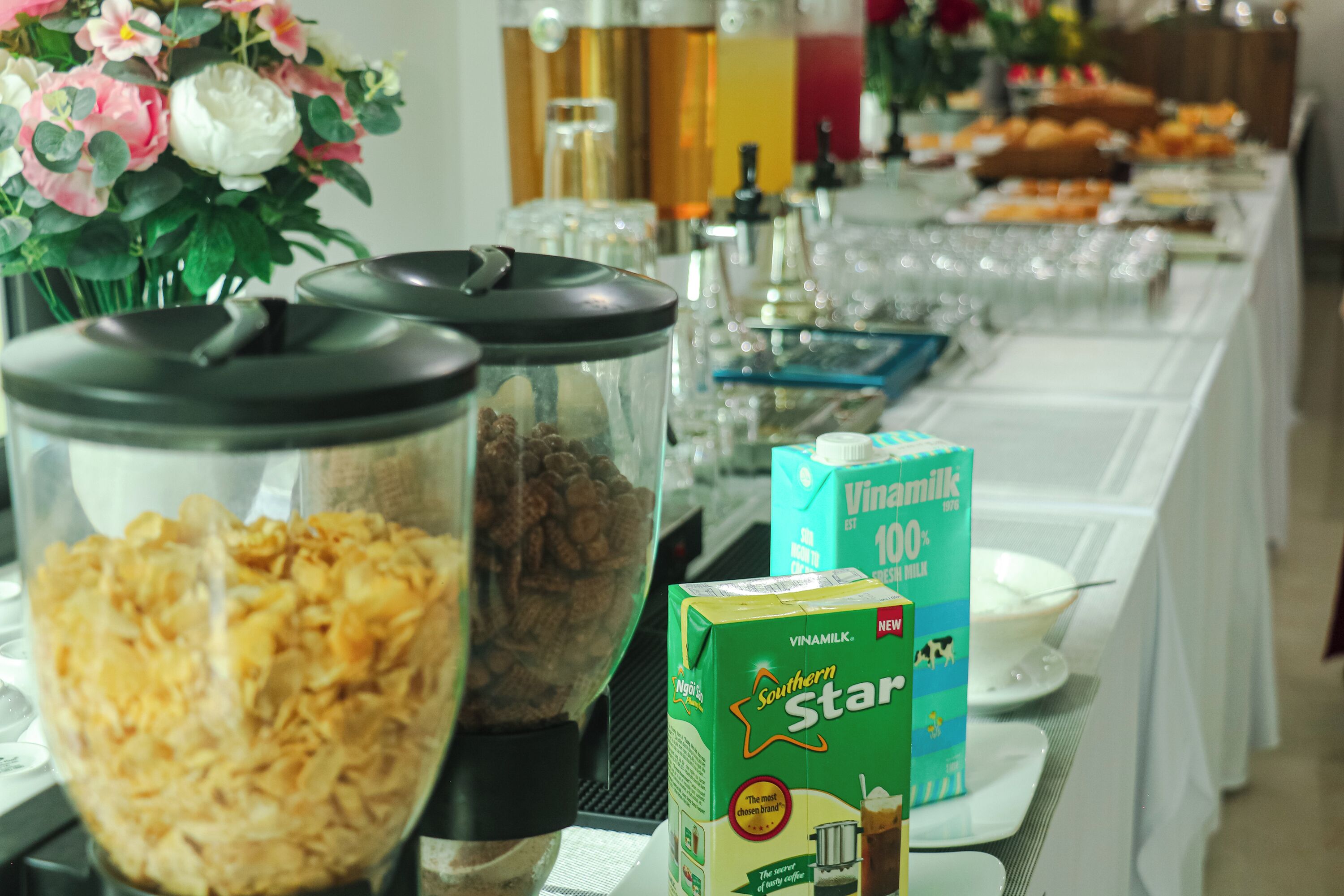 A buffet table with food containers, a box of Vinamilk 100% milk, and a floral arrangement.