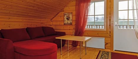 A cozy cabin interior with a wooden ceiling, a red sofa, a small table, and a window with a view of trees.