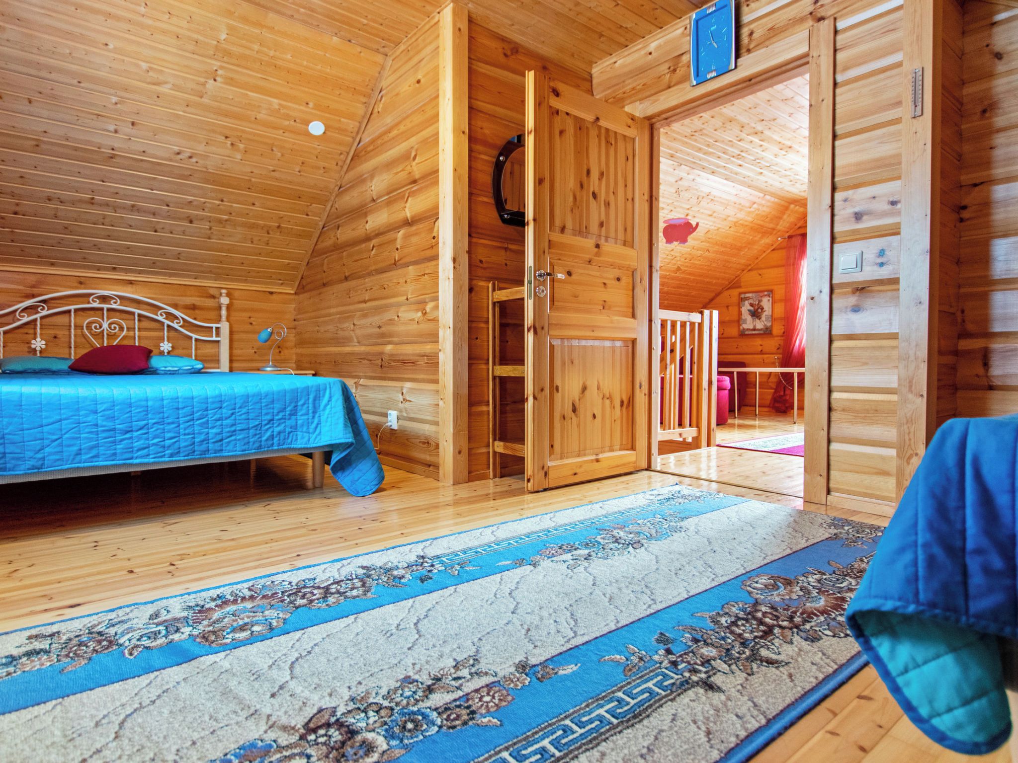 A wooden cabin interior with a bed, a door leading to another room, and a blue patterned rug.