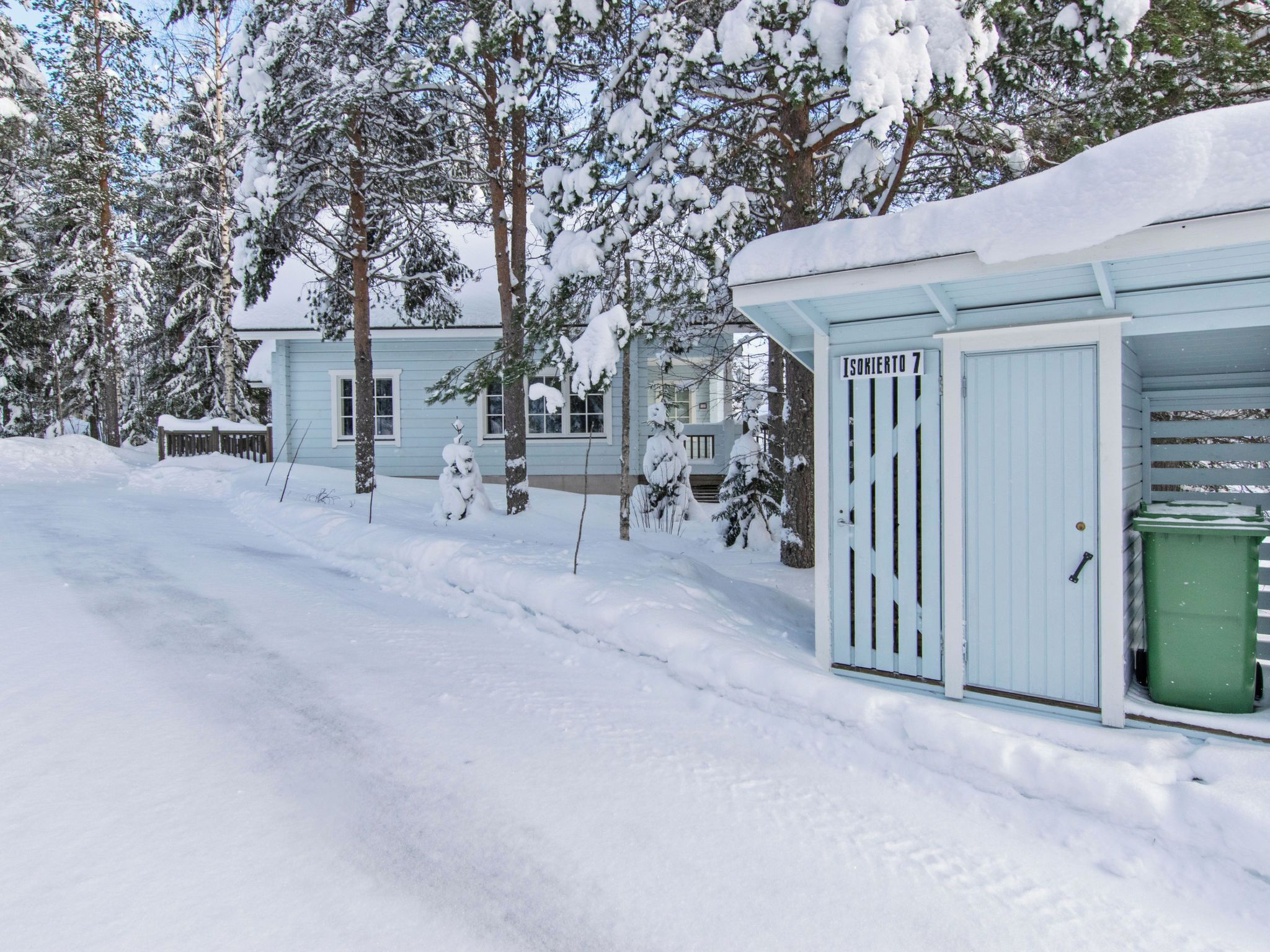 Een door de sneeuw bedekte locatie met een lichtblauw gebouw, een groene prullenbak en een bord waarop 'Isokierto 7' staat.