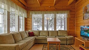 A cozy living room with a sectional sofa, a wooden coffee table, and a television on a wooden stand. Large windows with curtains allow natural light and views of a snowy landscape. - Lomataivas mökki by Interhome (Hyrynsalmi)