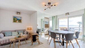 A modern living room with a sofa, coffee table, and dining area with chairs. There is a painting on the wall and a view of the sea through large windows. - Le Palazzo del Mar by Interhome (Cavalaire)