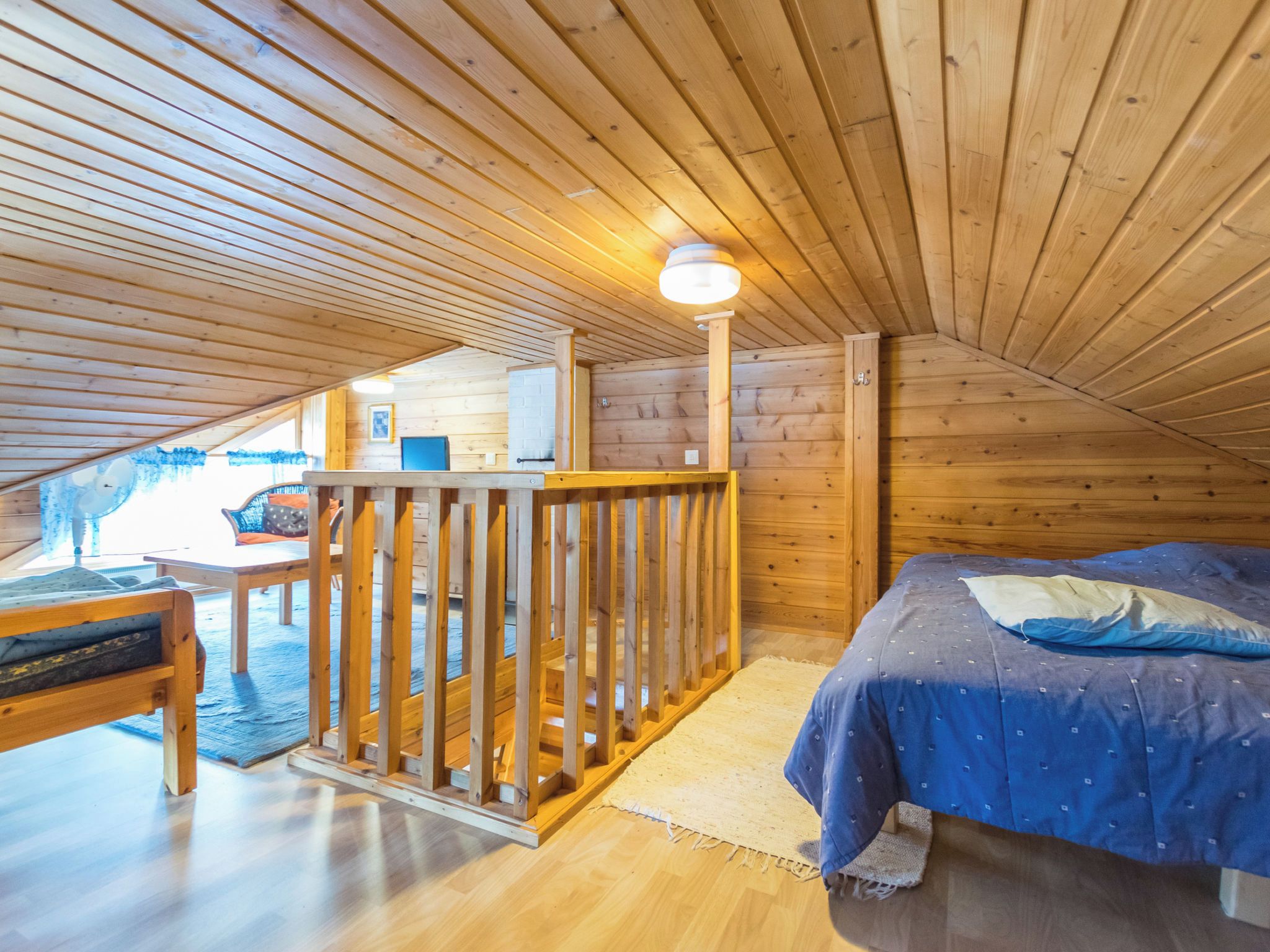 Une chambre mansardée chaleureuse avec un plafond en bois, un lit recouvert d’une courtepointe bleue, un bureau en bois et une petite table avec des chaises.