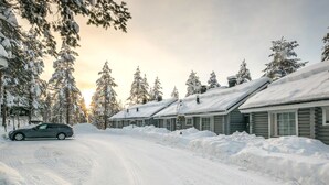A snow-covered road leading to a row of log cabins with a car parked on the left.