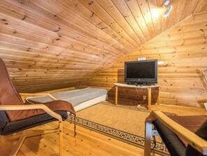 A small, cozy room with wooden walls and a sloped ceiling, featuring a single bed, a chair, a small table with a television, and a patterned rug.