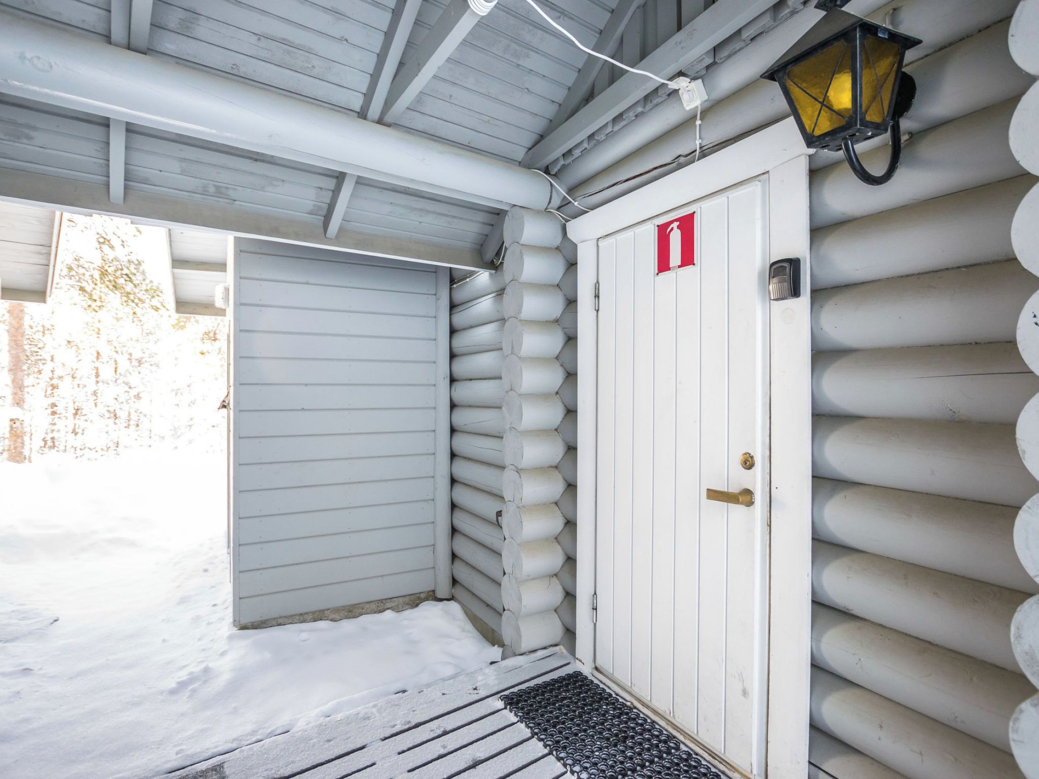 Une cabane en rondins avec un toit en métal, un panneau indiquant l’emplacement d’un extincteur et une lanterne fixée au mur.