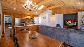Una cocina moderna con armarios de madera, un comedor con mesa de madera, y una sala de estar con sofá gris y televisor.