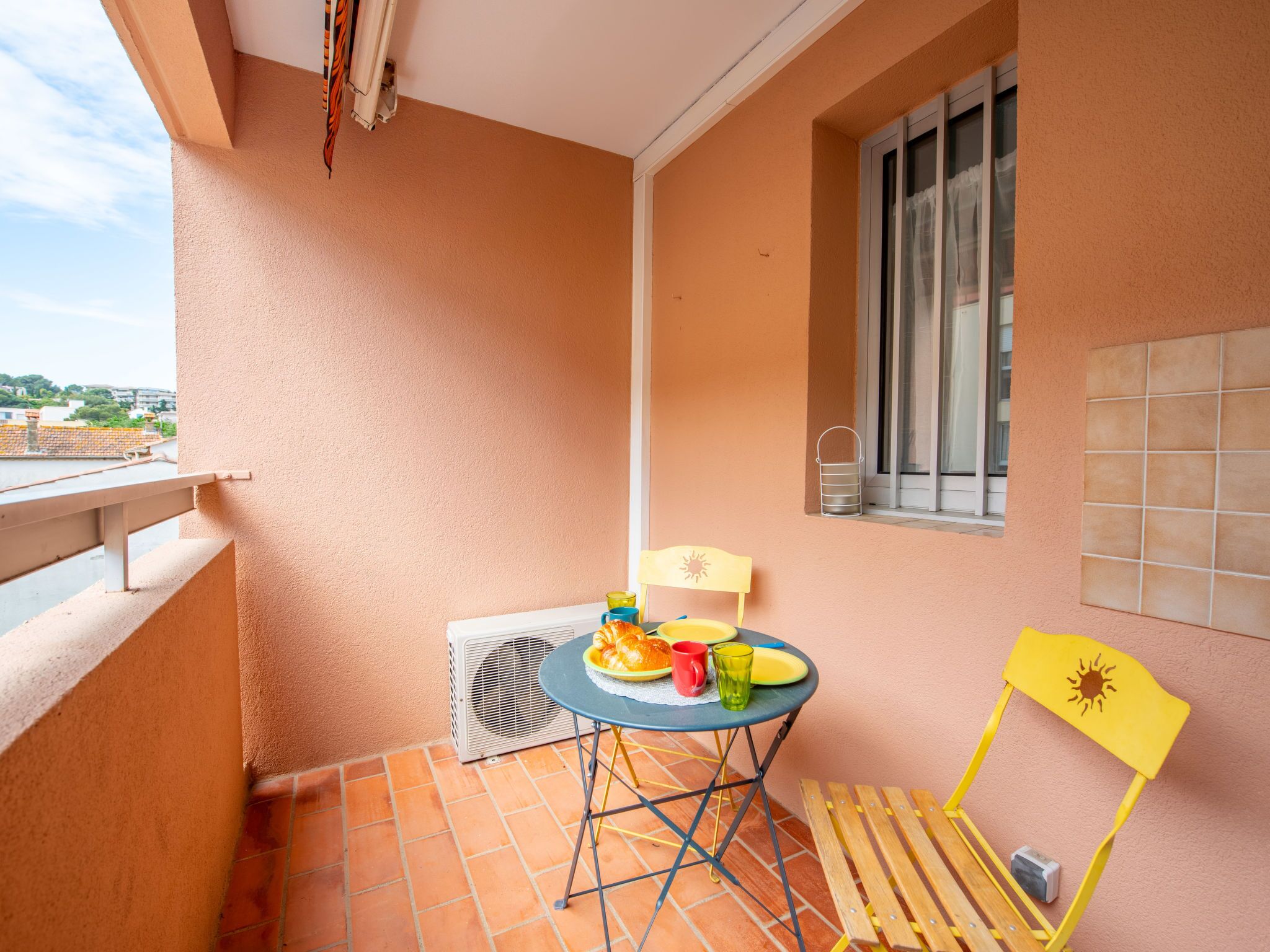 A balcony with a table set for breakfast, a chair, a window, and a view of the cityscape.