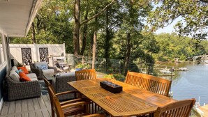 An outdoor deck with a wooden table and chairs, wicker sofas, and a glass railing overlooking a lake with boats.