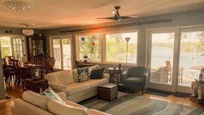 A spacious living room with a large window overlooking a body of water, a ceiling fan, and a dining area with wooden furniture.