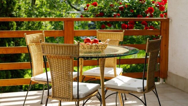Un balcon agrémenté de fauteuils en osier et d’une table en verre, entouré d’une végétation luxuriante et de fleurs rouges.