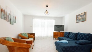 Un salon avec un canapé bleu, des fauteuils en osier, un téléviseur posé sur un meuble en bois et un tableau accroché au mur.