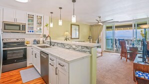 A modern kitchen with white cabinets, a microwave, an oven, and a dishwasher. There is a dining area with a table and chairs, and a view of the ocean through large windows. - "Oceanfront Condo: Coconut Tree & Ocean Views, Whale Watching, Paddlers, and Surfers at Wailua Bay!" (Kapaa)