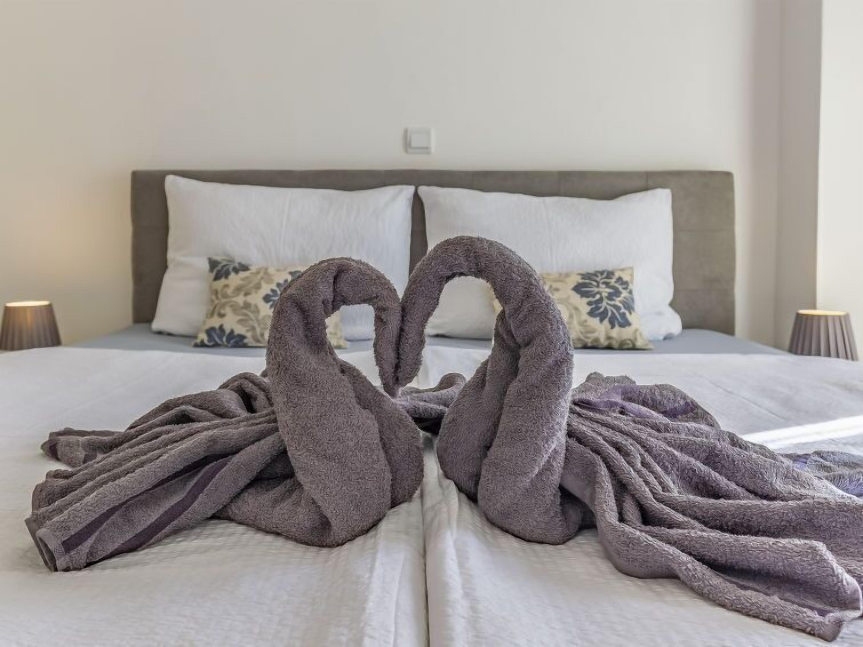 Una cama con un edredón gris y dos toallas dispuestas en forma de corazón.