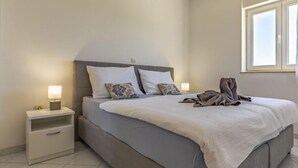 A neatly made bed with a grey headboard, a bedside table with a lamp, and a window with blinds.
