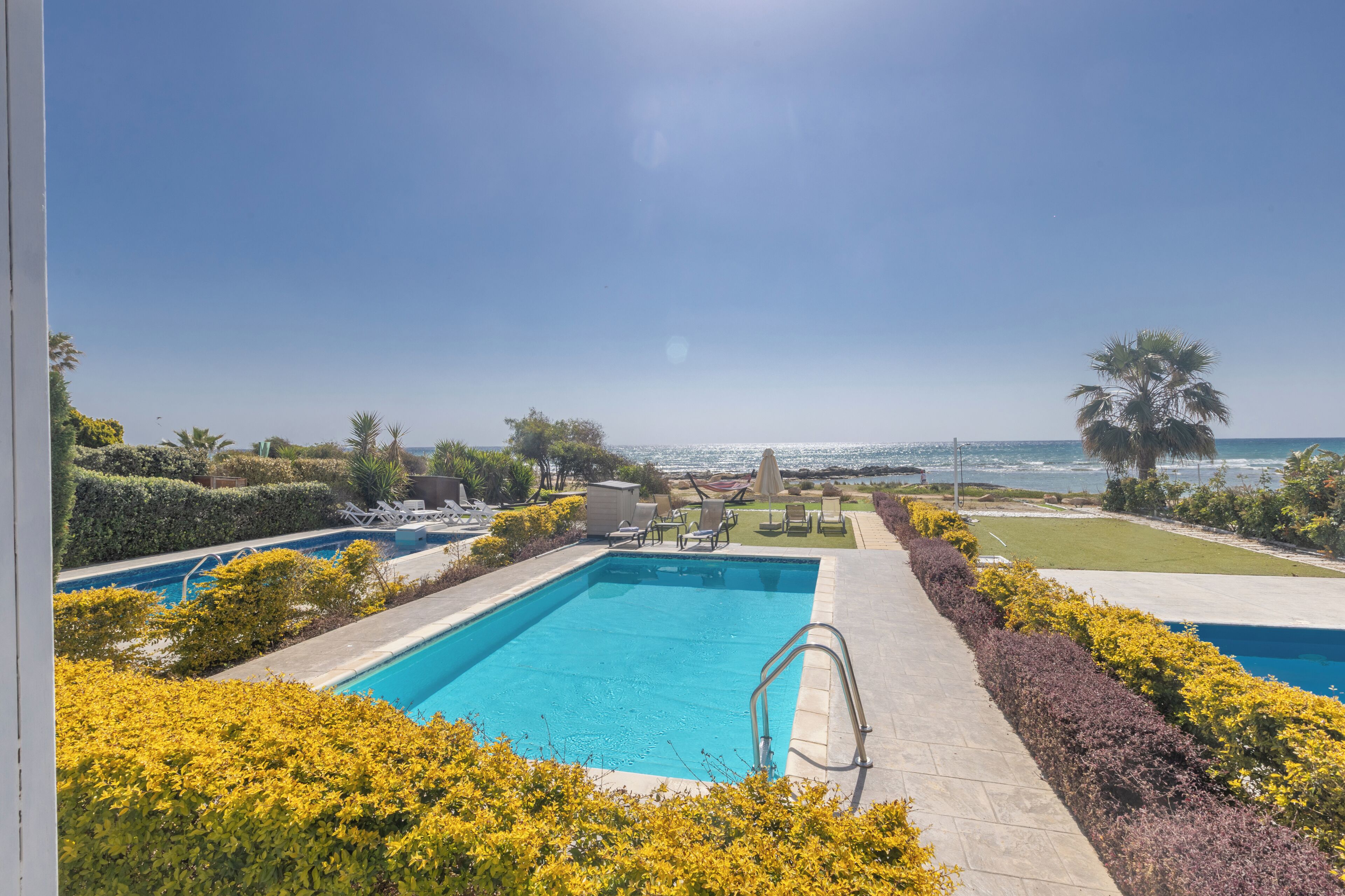 Une piscine avec vue sur l’océan, entourée de haies bien entretenues et sous un ciel dégagé.