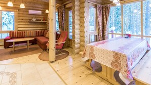 A cozy log cabin interior with a sofa, a chair, a table, and large windows offering a view of the forest. - Sateenkaari mökki by Interhome (Hyrynsalmi)