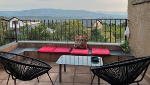 Een balkon met een tafel, twee stoelen, rode kussens en uitzicht op verre bergen.