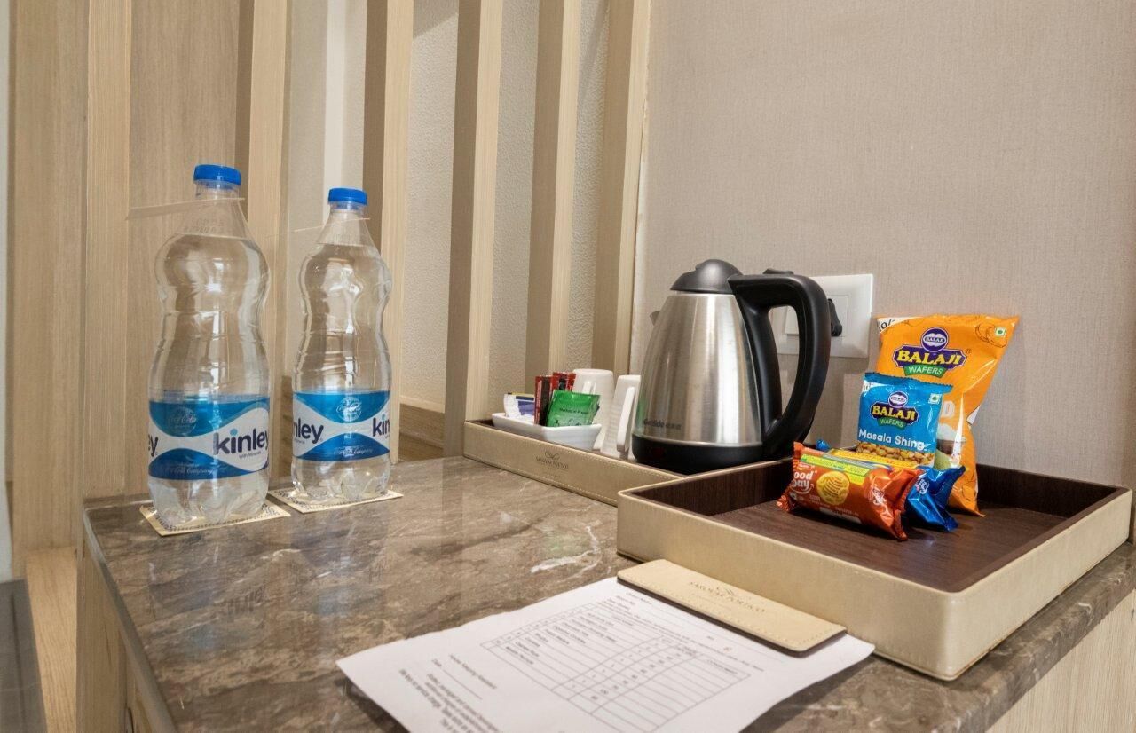 A hotel room counter with two water bottles, a kettle, a packet of Balaji Masala Shing, and a tray with a small dish and a cup.