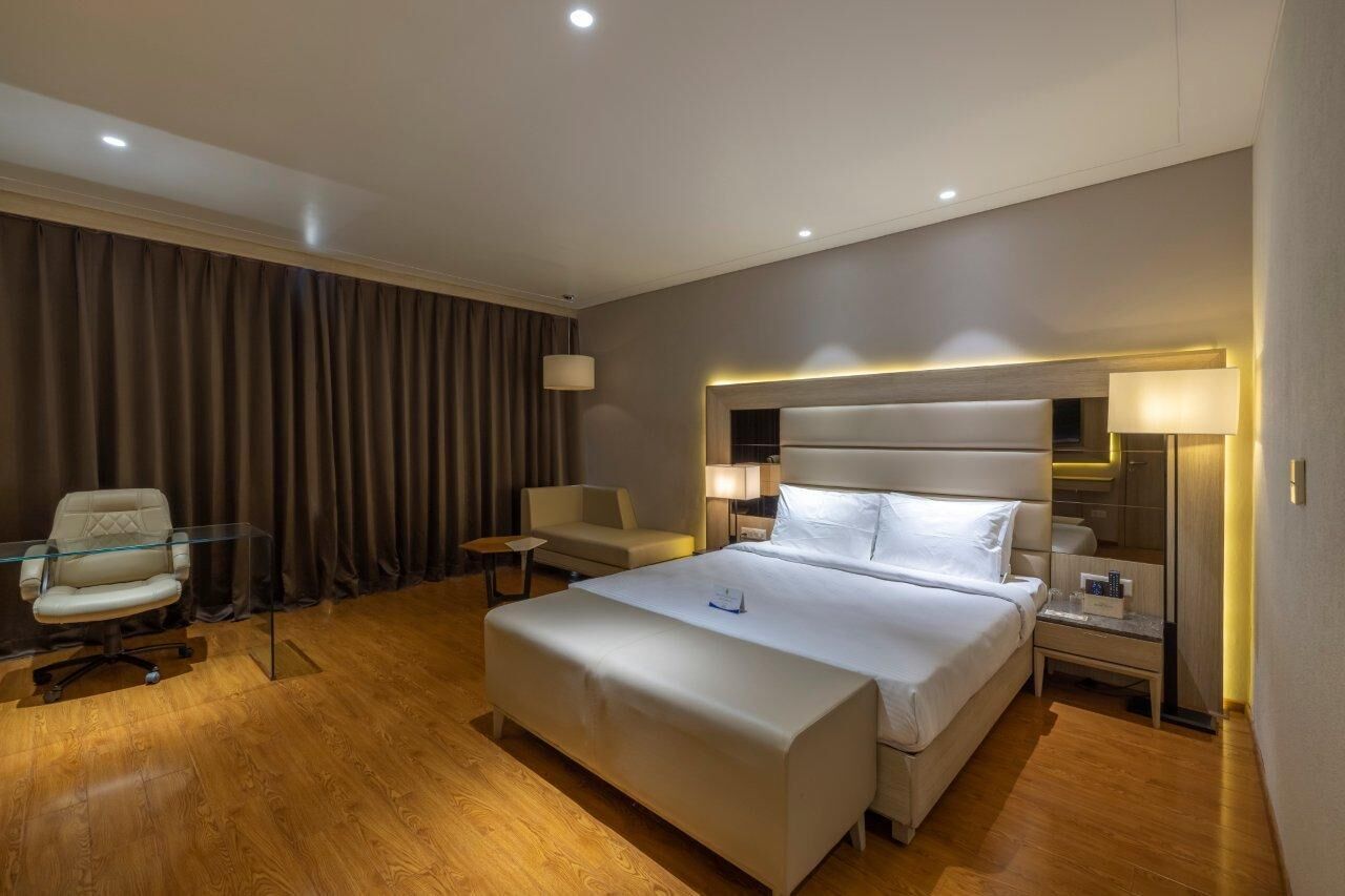 A modern hotel room with a large bed, a glass desk, a chair, and a bedside table with lamps.
