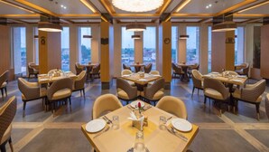 Un espace de restauration moderne, doté de plusieurs tables et chaises, de grandes fenêtres offrant une vue sur la ville et d’un lustre central.