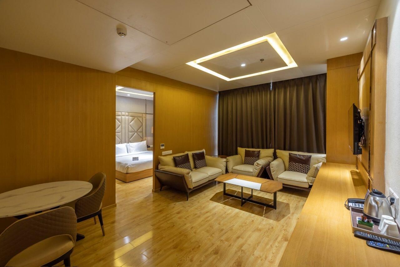 A modern hotel room with a wooden floor, a sofa set, a dining table, and a TV mounted on the wall.