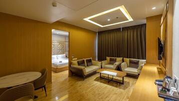 A modern hotel room with a wooden floor, a sofa set, a dining table, and a TV mounted on the wall.