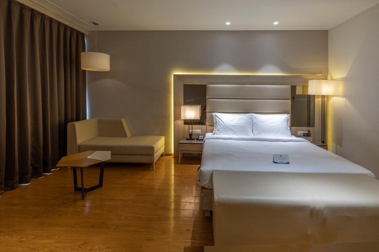 A hotel room with a bed, a chair, a small table, and a nightstand with a lamp.