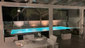 A modern outdoor pool area with glass walls, white furniture, and ambient lighting.