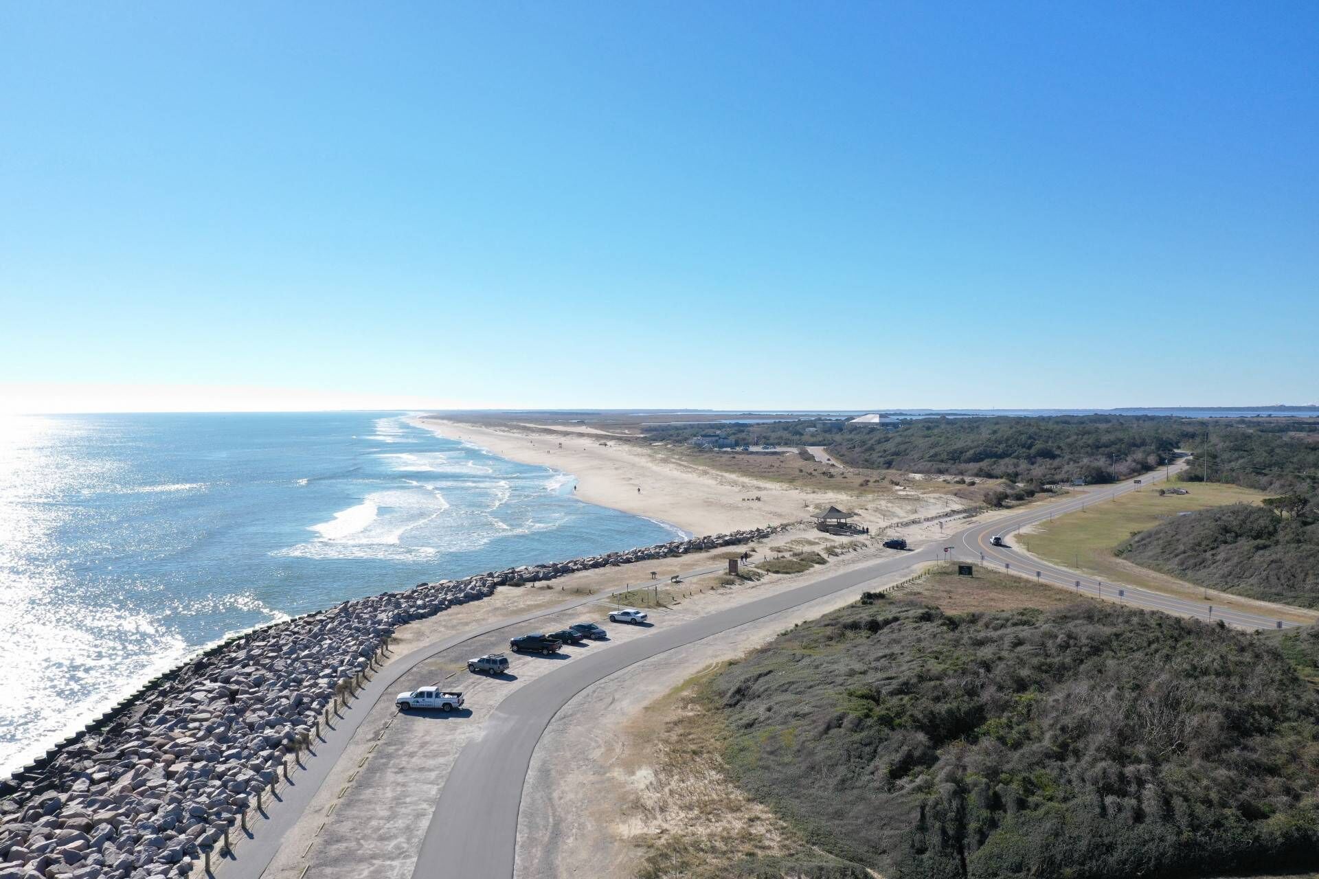 An aerial view of a coastal road with a beach on one side and a forested area on the other. There are several cars parked along the road, and a large number of sheep are gathered near the beach.