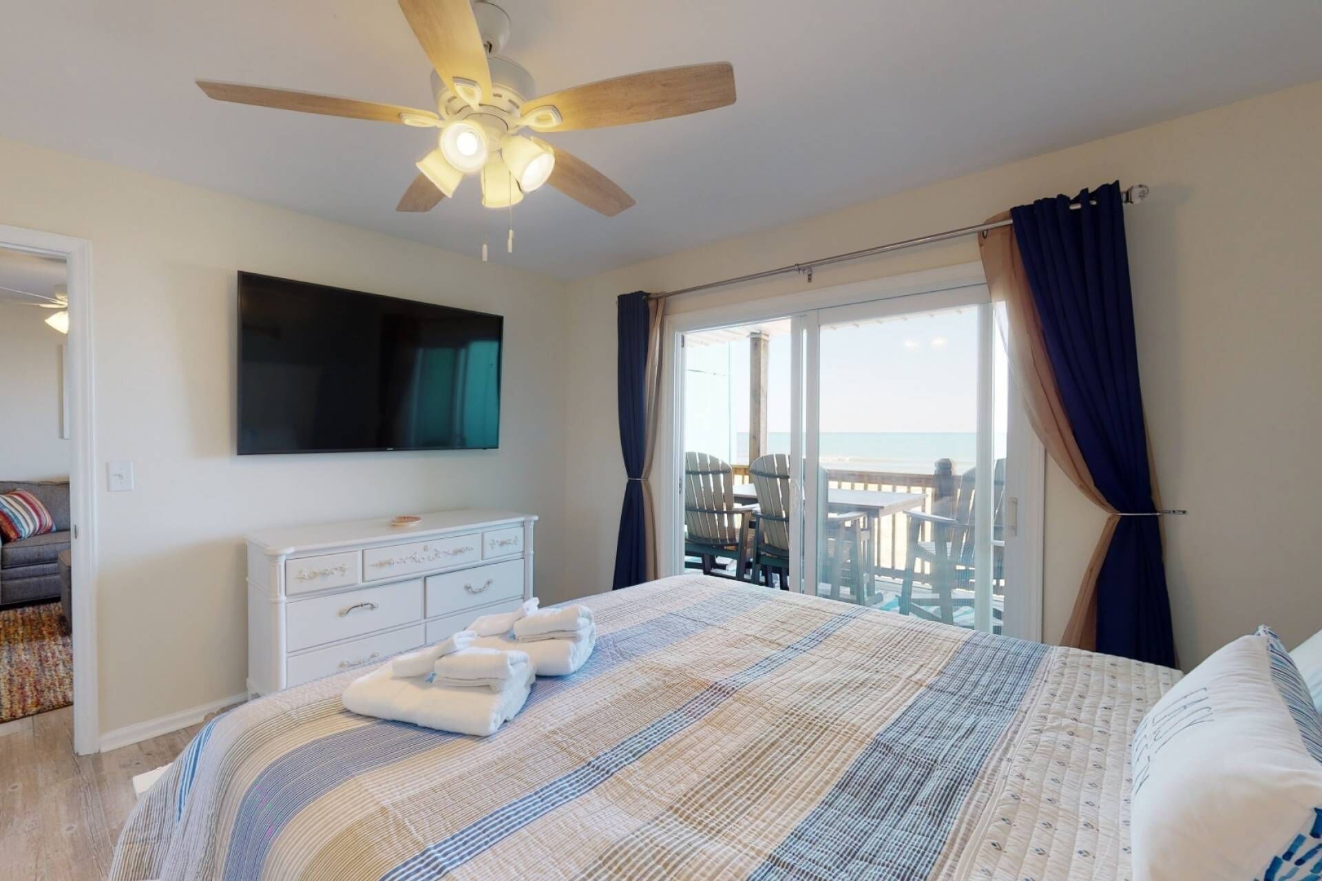 A bedroom with a bed, a TV, a ceiling fan, a dresser, and a balcony with a view of the sea.