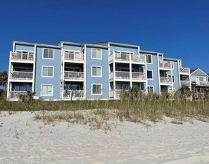 Un edificio de varios pisos frente a la playa, con balcones, probablemente un hotel o complejo de apartamentos.