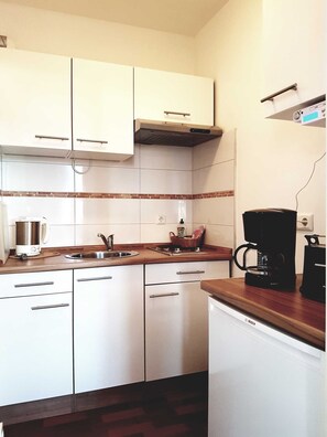 Cocina compacta con armarios blancos, un lavamanos de acero inoxidable, una cafetera Bosch y un refrigerador pequeño.
