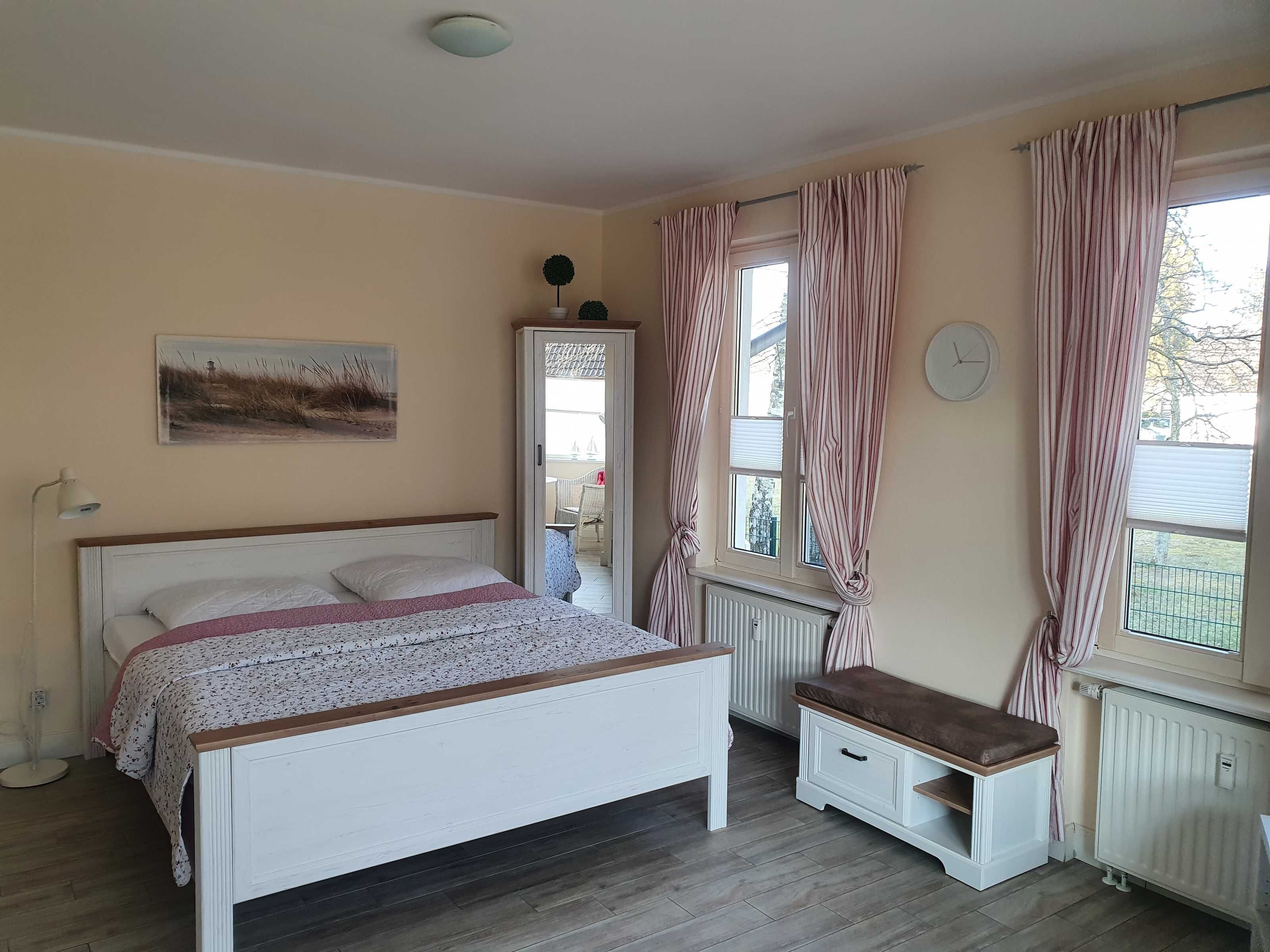 Quarto com cama, mesa de cabeceira, banquinho, radiador, janela com cortinas, relógio de parede e um quadro.