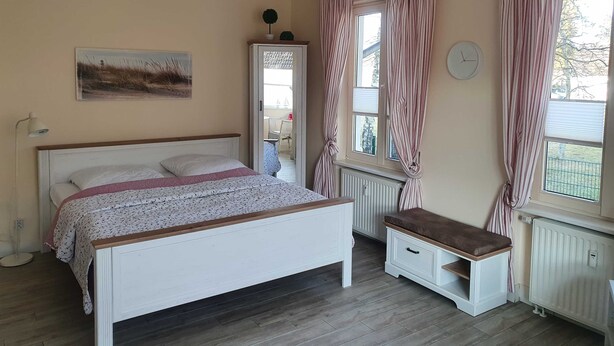 Un dormitorio con cama, mesita de noche, banco, radiador, ventana con cortinas, un reloj de pared y un cuadro.