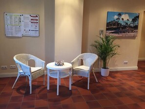 Una habitación con dos sillas de mimbre, una mesita redonda con un adorno, una planta en maceta, un tablón de anuncios y un cuadro de una escena de playa en la pared.