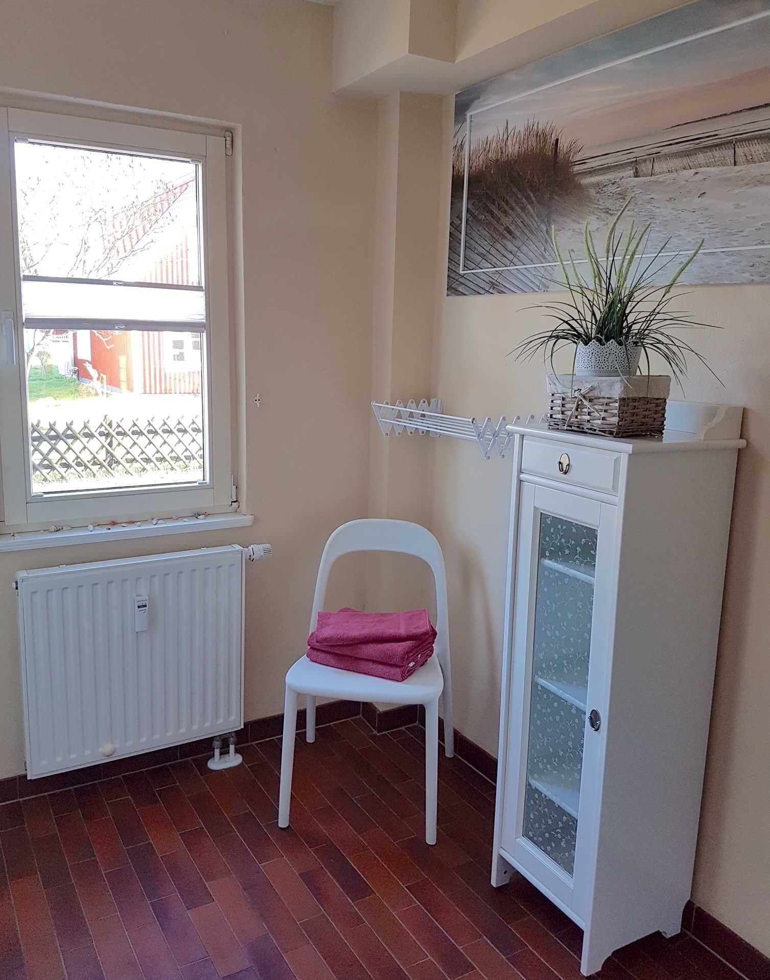 A room with a radiator, a window, a chair with a pink towel, a cabinet with a plant, and a wall-mounted artwork.