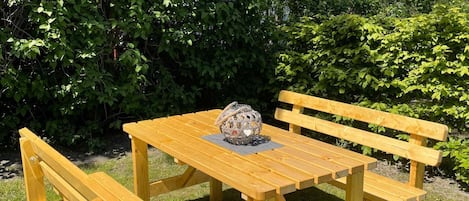 A wooden picnic table with benches, surrounded by lush greenery and a red brick house in the background.