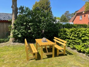 Een houten tafel en bankjes in een tuin met een boom en een haag.