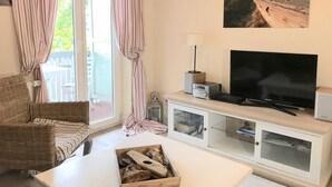 Un salon avec une télévision, un meuble blanc, un canapé en osier et une fenêtre avec des rideaux.