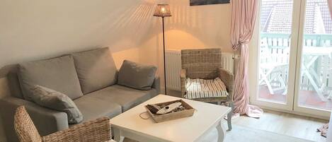 Een gezellige woonkamer met een bank, twee rieten stoelen, een kleine tafel, een vloerlamp en een raam met gordijnen.