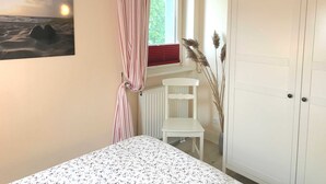 Ein Schlafzimmer mit Bett, einem Fenster mit Vorhängen, einem Heizkörper, einem weißen Stuhl und einem Kleiderschrank.
